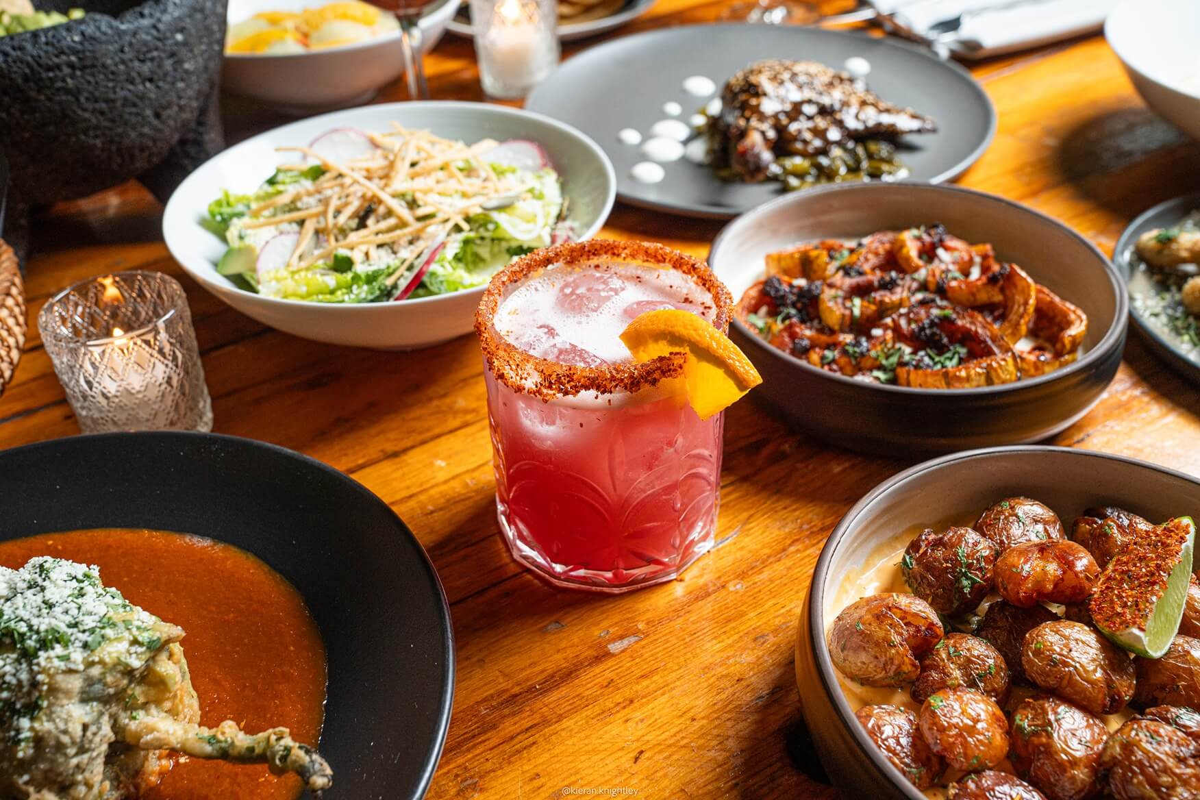 Table with various Mexican dishes including salad, roasted potatoes with lime, a sauced meat dish, and a pink cocktail with a chili-rimmed glass and orange slice.