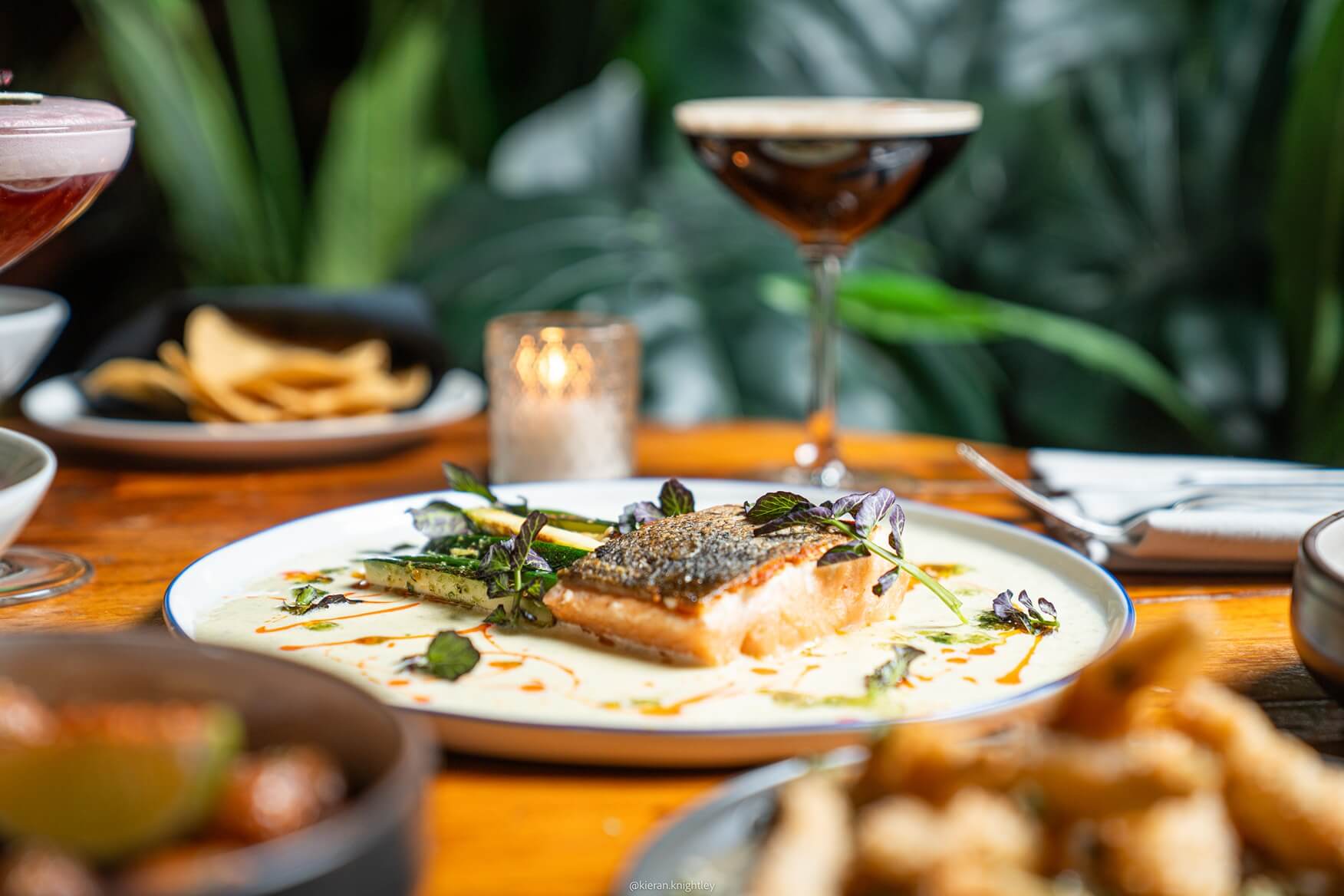 Plated salmon fillet with zucchini and herbs in creamy sauce on a wooden table with cocktails and chips in a restaurant setting.
