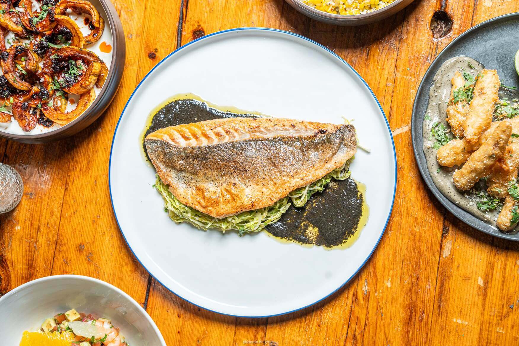Grilled fish fillet served on shredded greens with a dark sauce on a white plate on a wooden table surrounded by bowls of various side dishes.