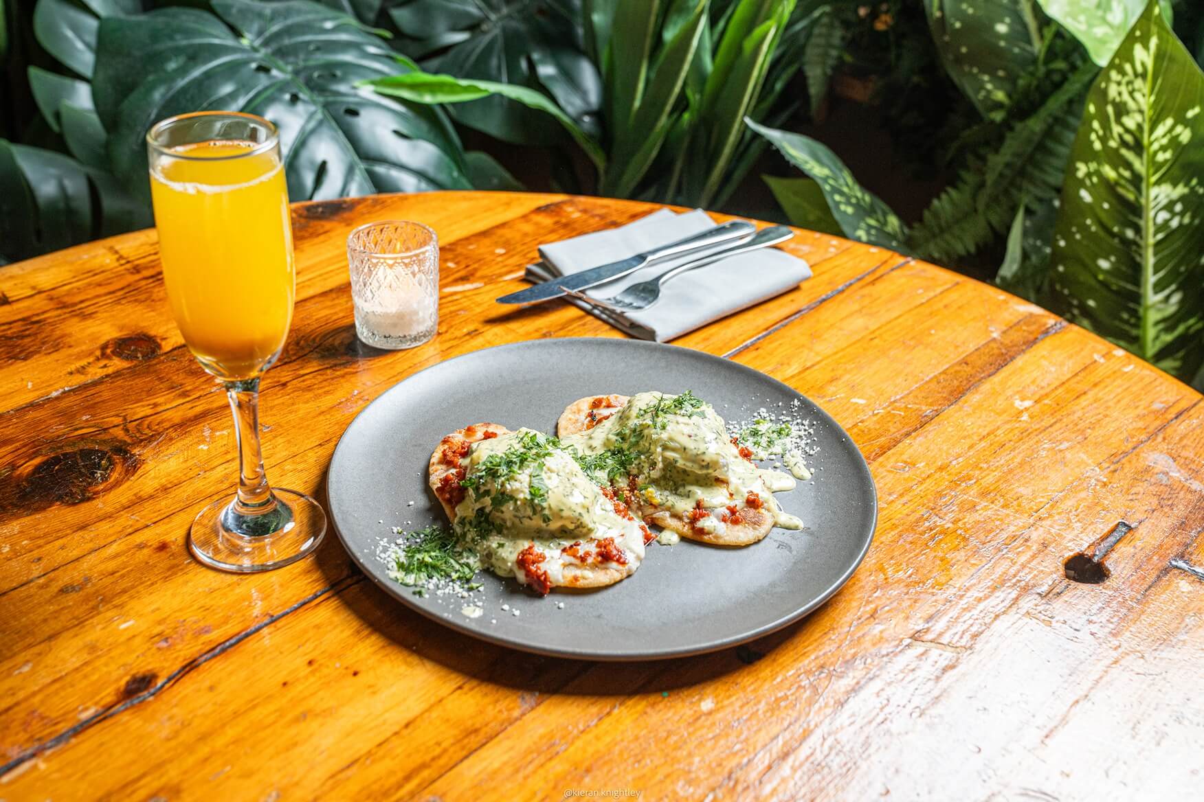 Plate of two small tortillas topped with creamy sauce, red salsa, and chopped herbs on a wooden table with a glass of orange juice and silverware in the background.