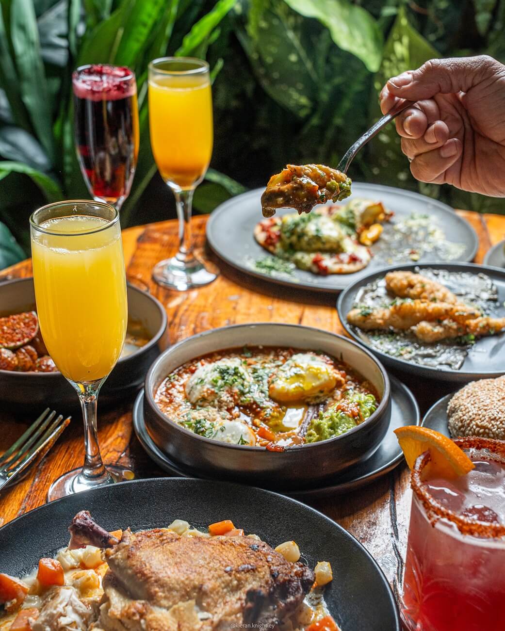 Table with various plated dishes including roasted meat, stewed vegetables, fried food, and four colorful drinks, with a hand holding a spoonful of food.