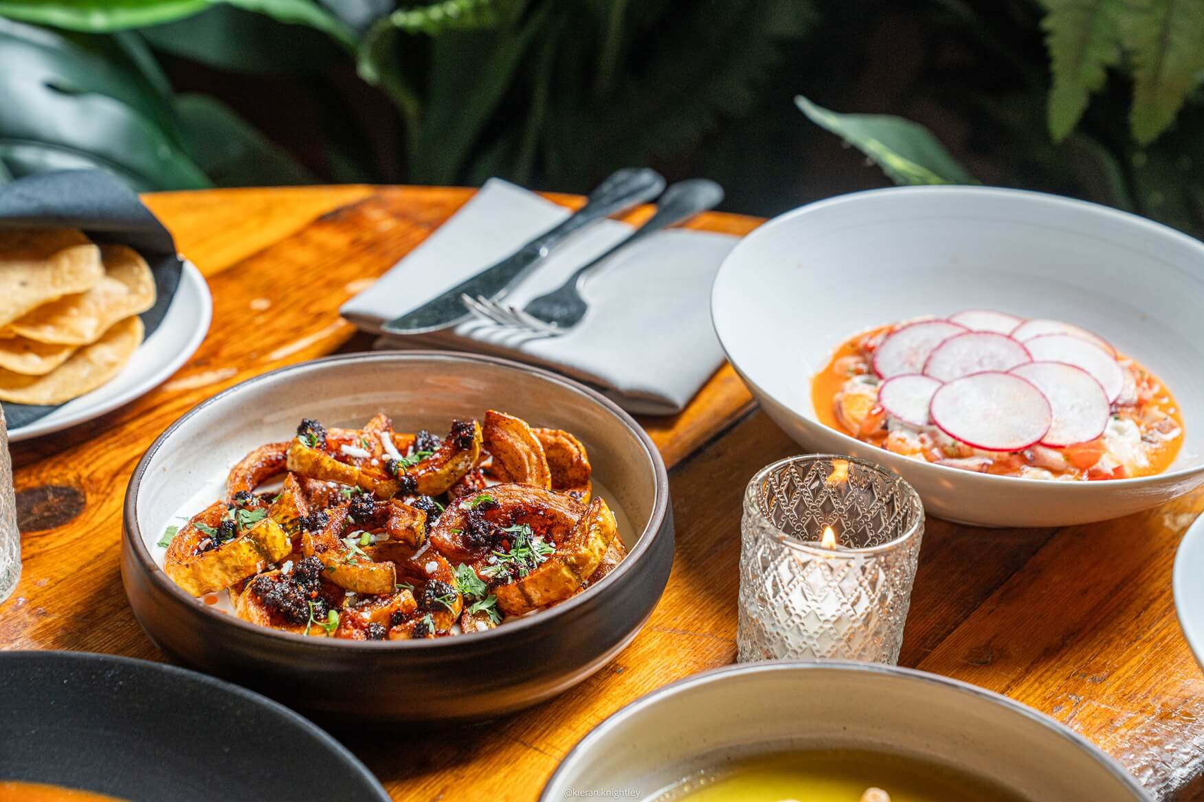 Wooden table with bowls of Mexican food including grilled squash with spices, a dish topped with sliced radishes, bowl of tortilla chips, and a lit decorative candle.