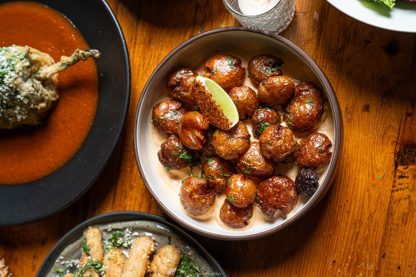 Bowl of crispy roasted baby potatoes garnished with herbs and a lime wedge, served on a creamy sauce on a wooden table with other dishes nearby.