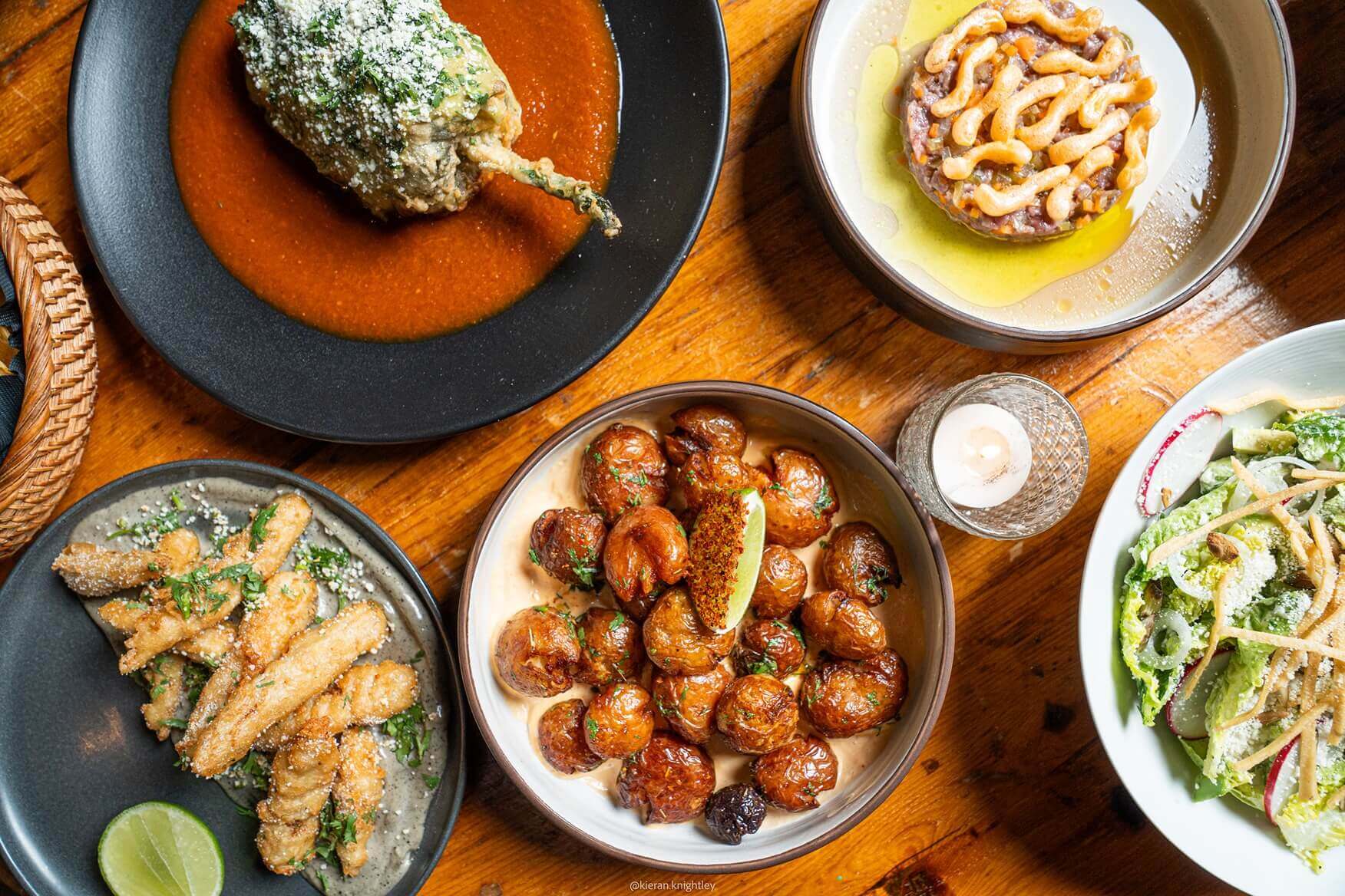 Top-down view of five dishes on a wooden table including fried stuffed pepper with red sauce, fried finger food with lime, roasted potatoes with a lime wedge, a salad with radishes and tortilla strips, and a plated dish topped with sauce.