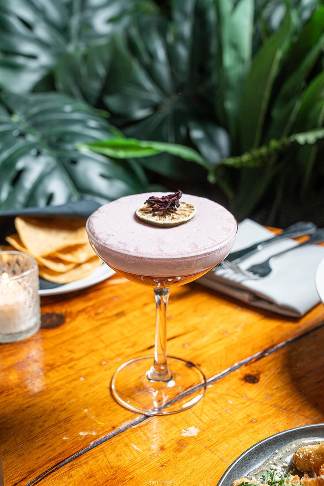 Foamy cocktail garnished with a dried citrus slice and flower petal on a wooden table with a plate of chips and a candle nearby.