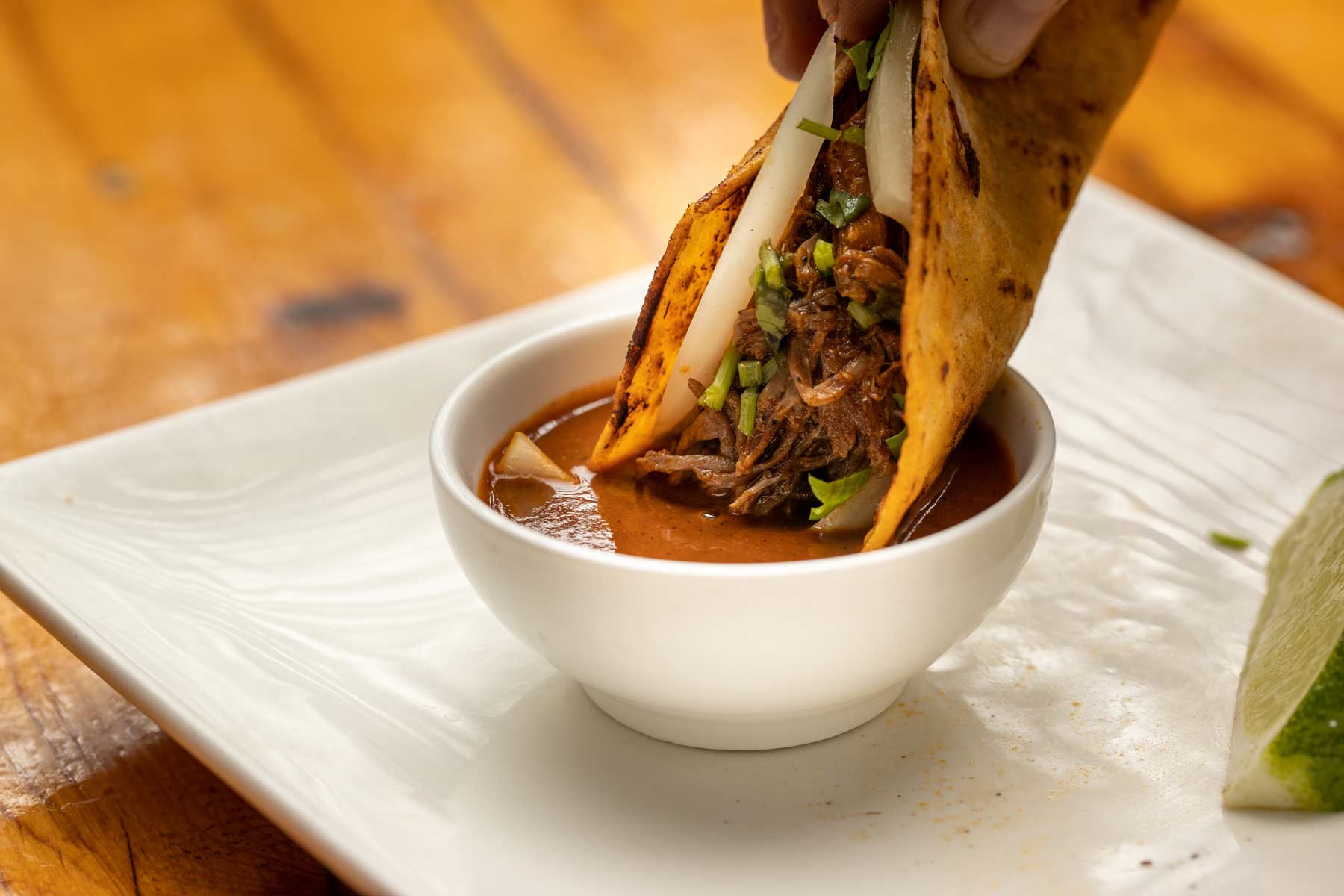 Hand dipping a taco filled with shredded meat, onions, and herbs into a bowl of red sauce on a white plate.