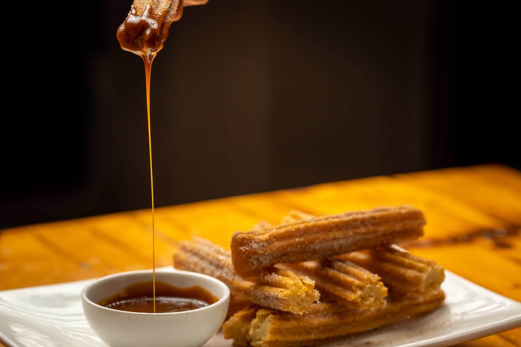 Caramel sauce being drizzled over a white bowl next to sugar-coated churros on a white plate.