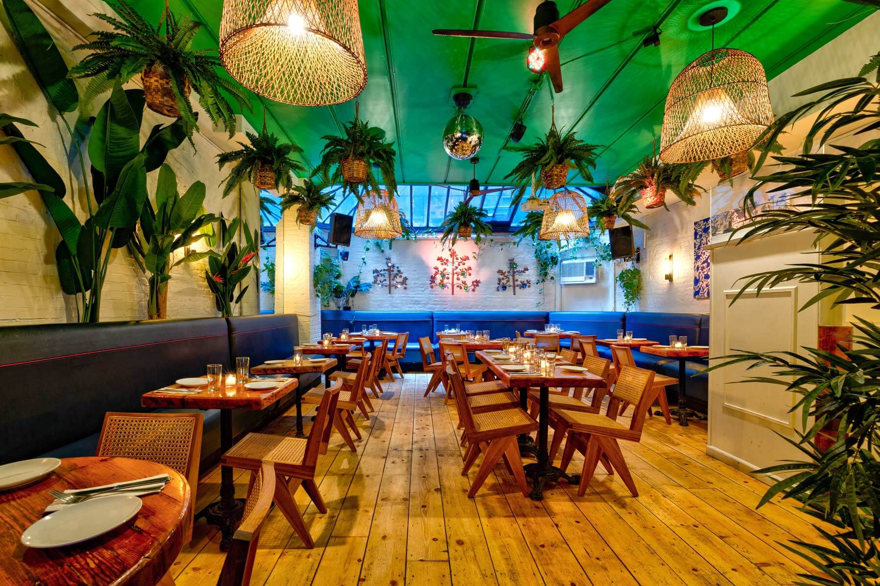 Cozy restaurant interior with wooden floors, hanging plants, wicker pendant lights, and wooden tables set with plates and glasses.