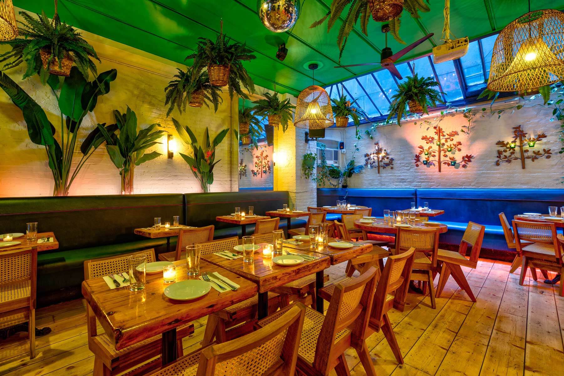 Cozy restaurant interior with wooden tables, rattan chairs, hanging plants, wicker light fixtures, and candles on tables.