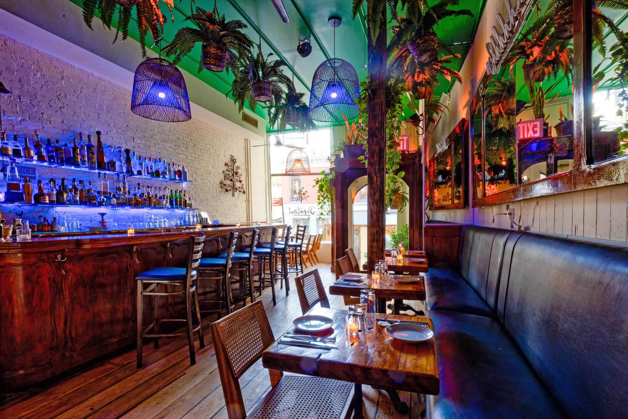 Cozy bar and dining area with wooden tables, chairs, a dark leather bench, hanging plants, and blue-lit shelves of bottles.
