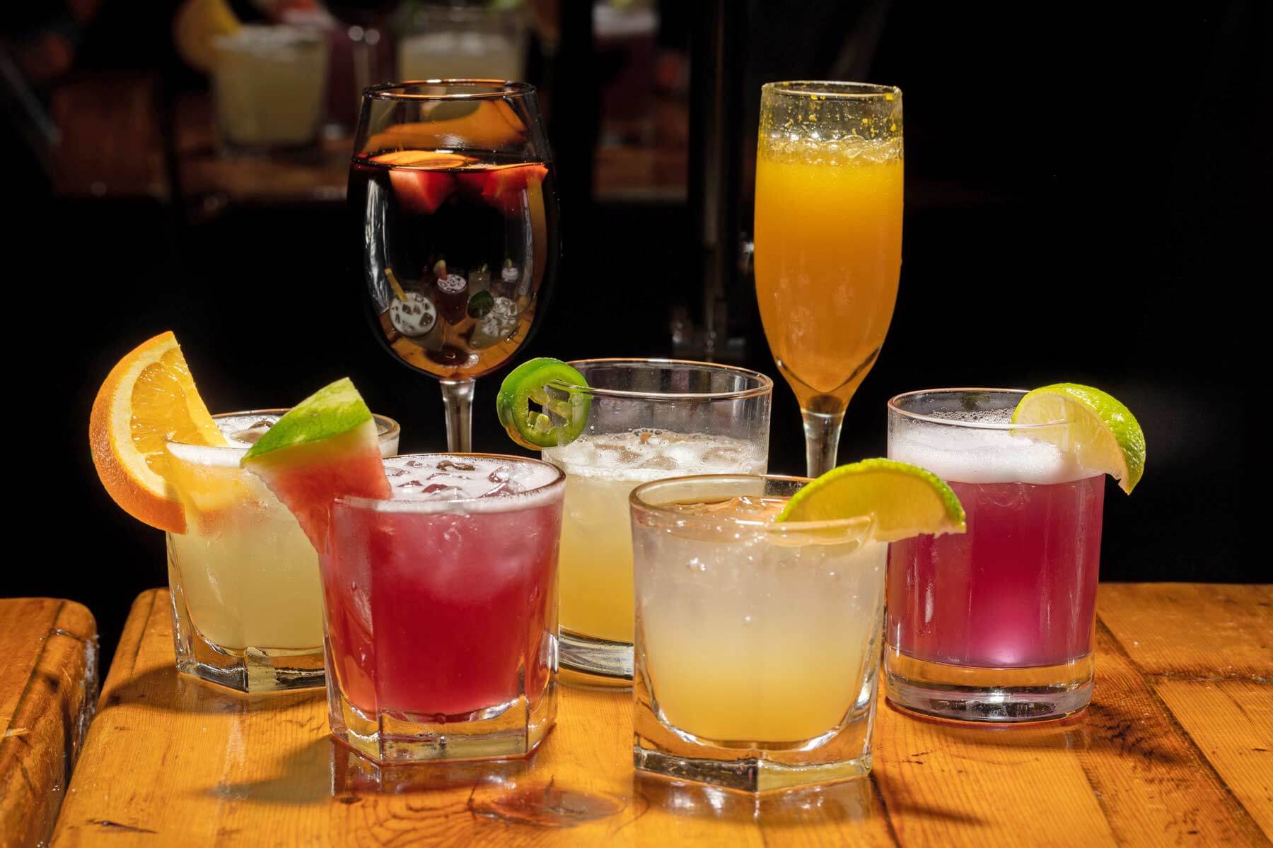 Seven colorful cocktails garnished with fruit slices and jalapeño, arranged on a wooden table against a black background.